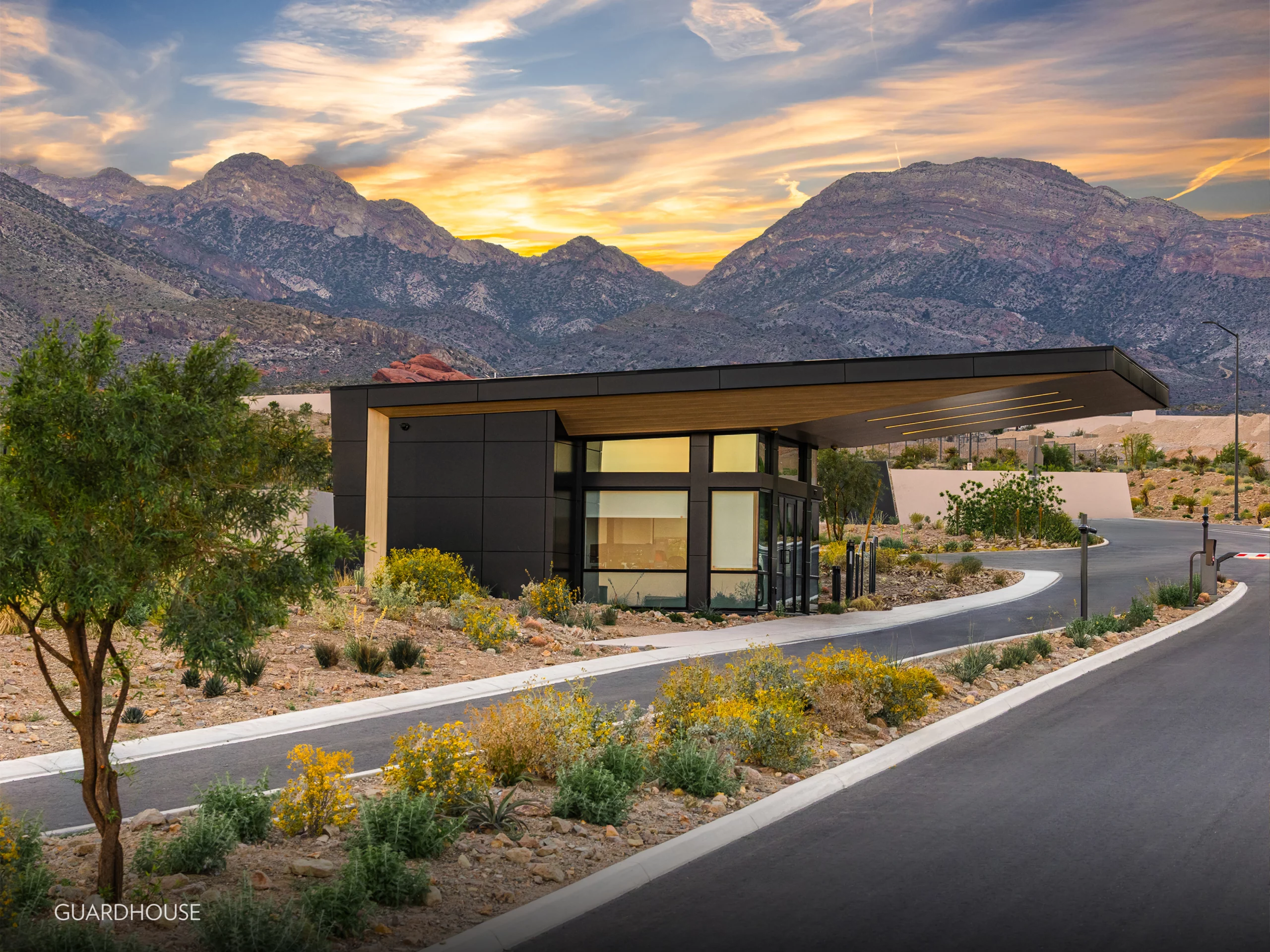 Astra_Guardhouse_NEW_1867x1400 A modern, custom guardhouse with large windows sits beside a curved road, surrounded by desert plants and southern mountains in the background under a colorful Las Vegas sunset sky.